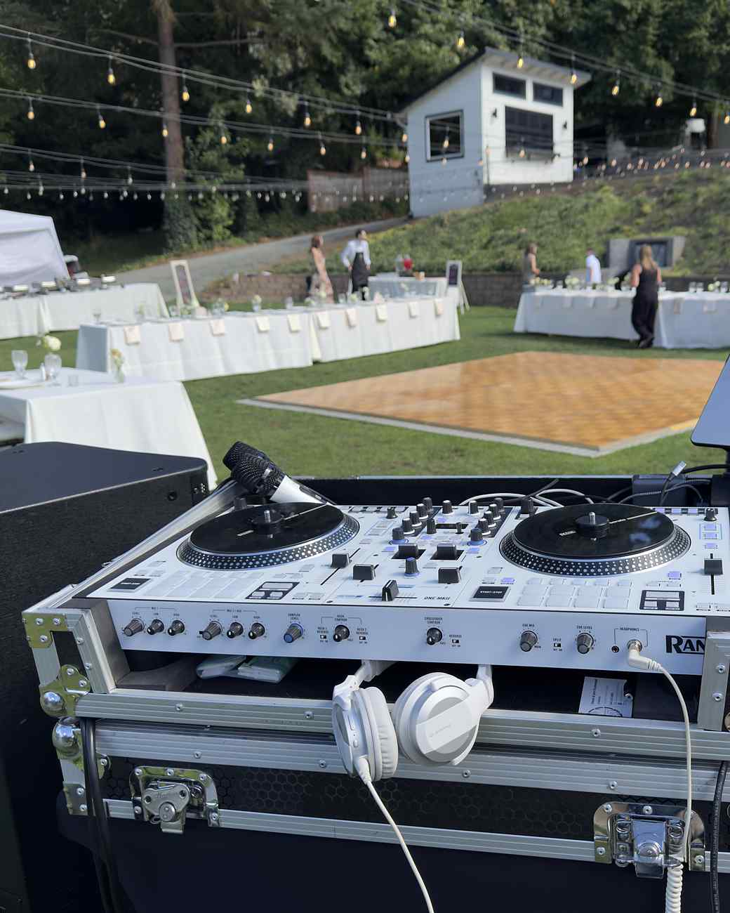 Two turntables either side of a mixer in an open road case, headphones hooked over the front edge, set up on the grass at an outdoor reception with the dance floor, string lights and laid tables behind.